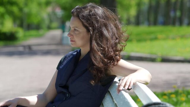 Beautiful brunette woman with long hair sitting on bench in public square on sunny spring day adult woman relaxes in park.