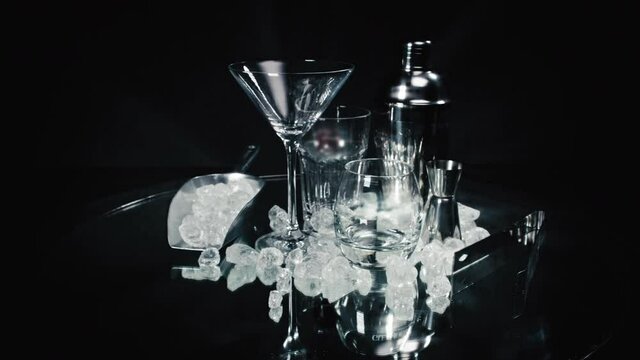 Camera moving away from a packshot of a cocktail shaker and glasses with ice cubes infront of a black background.