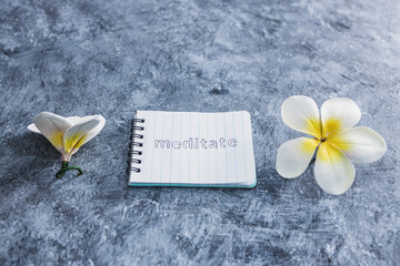 mental health concept, notepad with text Meditate next to tropical flowers on grey concrete surface