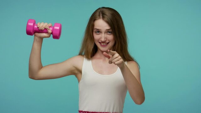 Cheerful lively girl holding small pink dumbbell and showing thumbs up, like gesture, recommending fitness training, maintain healthy sporty lifestyle. indoor studio shot isolated on blue background