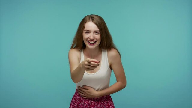 Amused happy girl pointing finger at camera and laughing out loud, holding belly with hilarious laughter, making fun of ridiculous appearance, joke. indoor studio shot isolated on blue background