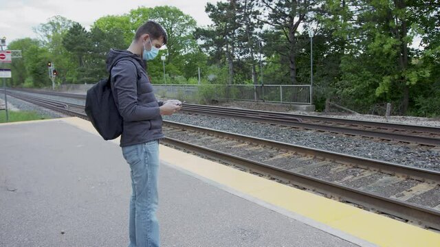 High quality 4k video of a young man who is waiting for a traing on a railway station. GO train station in Canada. Young handsome guy standing on a station and holding his cellphone wearing face mask