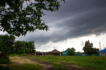 Fototapeta premium Heavy stormy gray sky. Waiting for the rain. Early spring in the village. Green vegetation, trees, bushes, grass. Houses are visible in the distance.