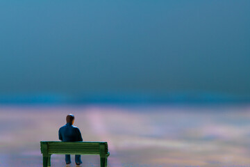 Miniature figurine posed as man sitting alone on a bench in surreal scenery, minimalist abstract concept image
