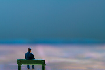 Miniature figurine posed as man sitting alone on a bench in surreal scenery, minimalist abstract...