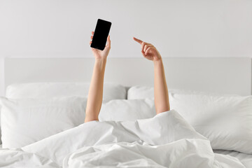 technology, comfort and morning concept - hands of young woman lying in bed with smartphone at bedroom