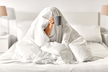 morning, comfort and people concept - young woman with cup of coffee sitting in bed under blanket...