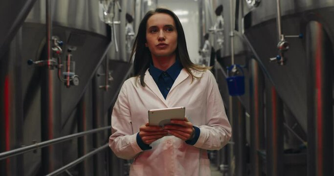 Scared female worker in white lab coat holding tablet and walking through rows of steel brewing vats. Alert signals and red lights at brewery factory. Concept of emergency situation