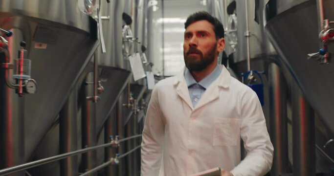 Male worker in white lab coat standing and using tablet while started running through rows of steel brewing vats. Alert signals and red lights at brewery factory. Emergency situation