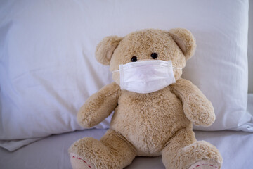 The teddy bear is sitting on a bed wearing a mask to protect against germs and viruses. Hygiene for children concept