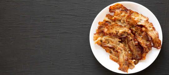 Homemade Fried Bacon on a white plate on a black surface, top view. Overhead, from above, flat lay. Space for text.