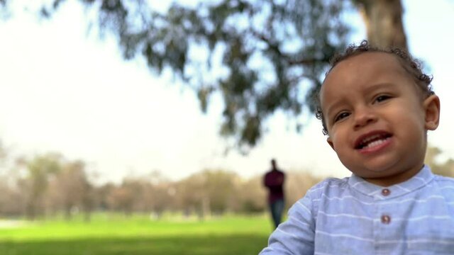 Little African American boy walking on lawn, holding red apple, biting and eating it. His father watching son out-of-focus. Family and weekend concept