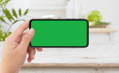 Left hand holding white mobile smartphone device green screen in kitchen