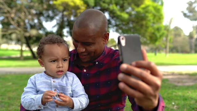 African American bald father trying to make selfie with cute son. Dad sitting on lawn in park, holding little boy and talking with him. Boy frowning and looking away. Family and weekend concept