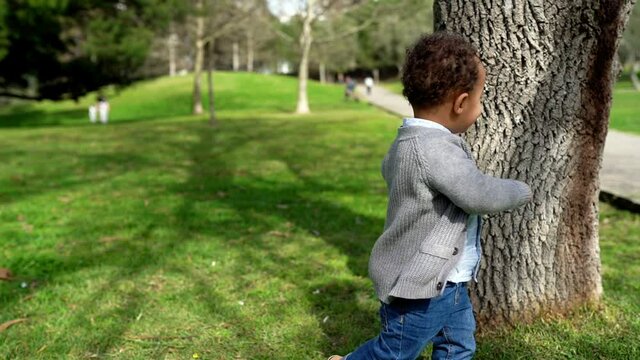 Happy African American little boy running around tree. Cute toddler playing hide-and-seek in city park at summer. Childhood and weekend concept