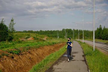 Obraz premium Stay in fit during quarantine. A sportive young woman is jogging outdoor, she have protective mask on face. Running alone on a tree-lined road in the days of the Corona Virus or Covid-19.