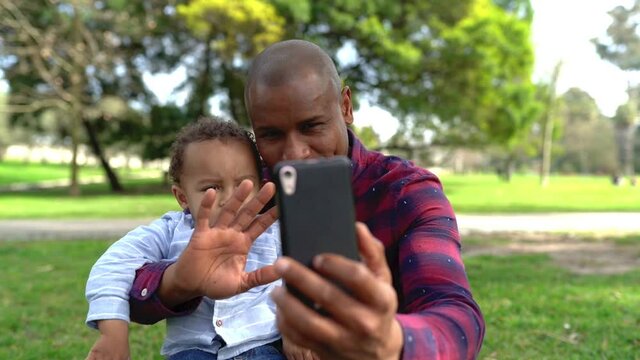 African American father waving and making selfie with son. Bald dad sitting on lawn, holding little boy and talking with him. Boy smiling and looking on father hand. Family and weekend concept