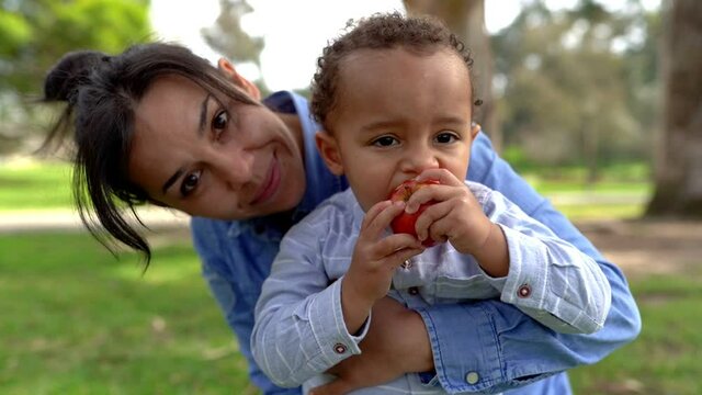 Little boy eating red apple, chewing and sitting on mom knees. Mixed-race young mother talking with him in city park. Childhood and weekend concept