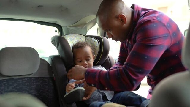 African American bald dad putting son into child car seat and securing him with belts. Little mixed-race boy ready for travelling in car. Safety and transportation concept
