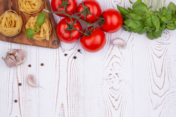 Top view raw tagliatelle pasta with fresh basil, garlic and tomatoes on a rustic white table, flat lay, copy space.