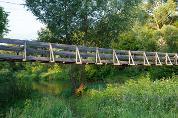 Beautiful summer day shot of an old narrow wooden hanging bridge over Nerl river with high green grass, vibrant vegetation, tall trees with rich foliage around. Ivanovo, Vladimir Oblast, Russia