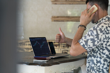 Satisfied man sits at home at a wooden table with laptop, talking on smartphone and is engaged in trading