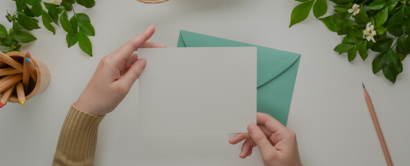 Female hands holding mock-up greeting card above turquoise envelope on white desk with decorations © bongkarn