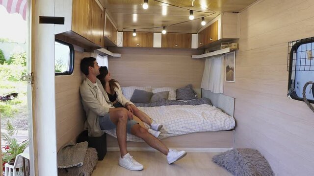 Well designed wheels house - smiling optimistic couple man and woman smiling while hugging in trailer mobile home, sitting on the bed