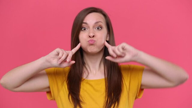Fun young woman in yellow t-shirt posing isolated on pink background studio. People lifestyle concept. Looking camera fooling around pointing index fingers on blowing cheeks monkey ears showing tongue