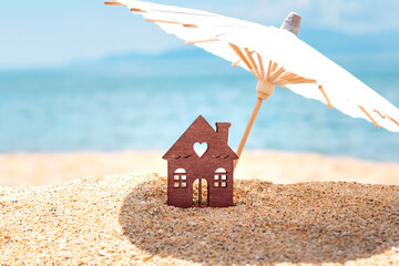 Miniature house and umbrella on beach, blue sea and sky on blurred background. Vacation home for...