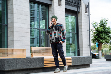 a young white man with a phone in his hands is sitting on a bench in an expensive neighborhood, a hipster in a hat is enjoying life, city streets, ethno clothing, hoodie