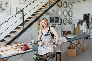 Portrait of smiling mature artist sitting in art studio with pictures and drinking coffee