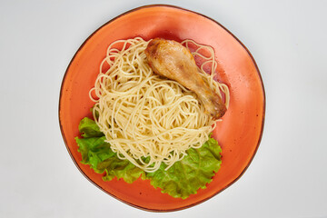 Spaghetti and fried chicken leg with a leaf of green salad lies on an orange plate. White background.