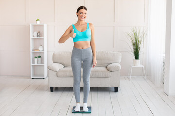Fit Girl Gesturing Thumbs-Up Standing On Weight Scales At Home