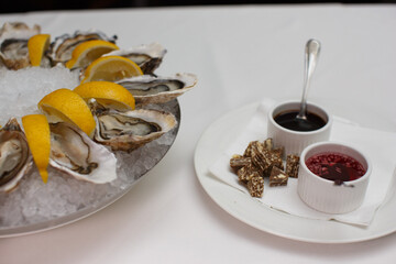 top view close up filled frame food shot of fresh raw shucked open oysters lying between lemon slices on a round cold ice tray next to Mignonette shallot vinegar and Ponzu sauces on a white table