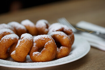 a pile of delicious sweet golden brown doughnuts (donuts) with white powdered icing sugar topping laying in a white plate on a wooden table background next to a fork and knife