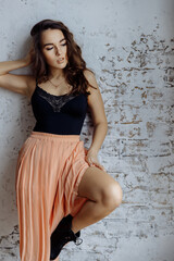 Fototapeta premium A beautiful brunette girl with curly hair stands against the wall in the loft style. The girl is wearing a light black top and skirt with pleating. High quality photo