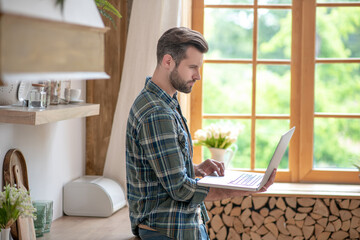 Man in a checkered shirt standing in the kitchen and holding a laptop in his hands, looking attentive