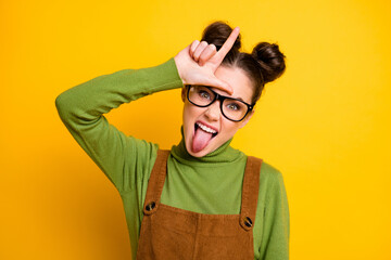 Photo of youngster student lady two buns bad person grimacing show loser symbol forehead bully classmate stick tongue wear specs green pullover brown overall isolated yellow color background - obrazy, fototapety, plakaty