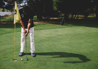Full length portrait of handsome golfer man in white pants and black t-shirt standing on golf course preparing to hit golf ball, good golf game at sunny day