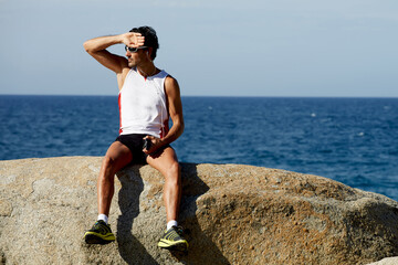 Handsome male jogger listening to music in headphones and enjoying seashore landscape in sunny day, mature sports man taking break after active training outdoors while holding smart phone in the hand