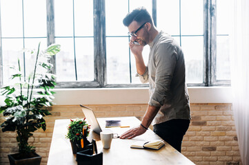 A side midshot of a young man that talks on the phone in the office, he is looking into computer monitor and typing with one hand. Young man wears casual clothes and eyeglasses