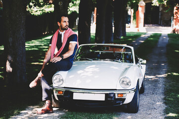 Handsome male looks wonderful sitting on the hood of luxury cabriolet car against his villa at...