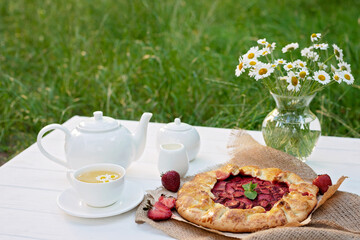 Freshly baked homemade galette or open strawberry pie, a cup of herbal tea, a teapot and a vase with a bouquet of daisy flowers, summer outdoor food. Soft selective focus.