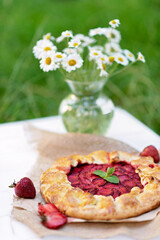 Freshly baked homemade galette or open strawberry pie and fresh mint leaves and a vase with a bouquet of daisy flowers at outdoor, summer food. Soft selective focus.