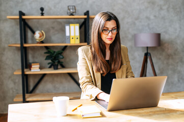 Young business woman works on a laptop in cosy office in casual smart wear, she sits at the table in eyeglasses and typing on a keyboard
