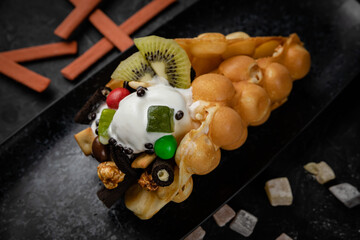 Hong Kong or bubble waffle with ice cream, fruits and colorful candies  on a dark background 
