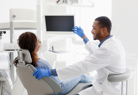 African man dentist talking to patient, pointing at blank monitor