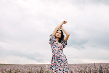 Young beautiful woman wearing a floral dress, standing in an endless field with pink wildflowers, on a blue sky background.