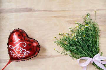 Bouquet of tiny white camomiles with green stems tied with pink ribbon and red heart-shaped balloon on wooden table. Ecological texture background for valentines birthday wishes with 